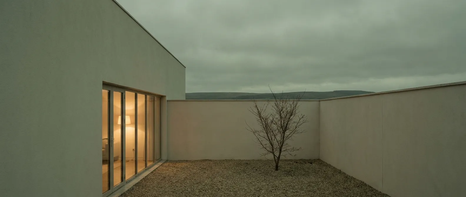 Walled gravel courtyard with a single bare tree, lamplight spilling from full-height glazing across the court at dusk