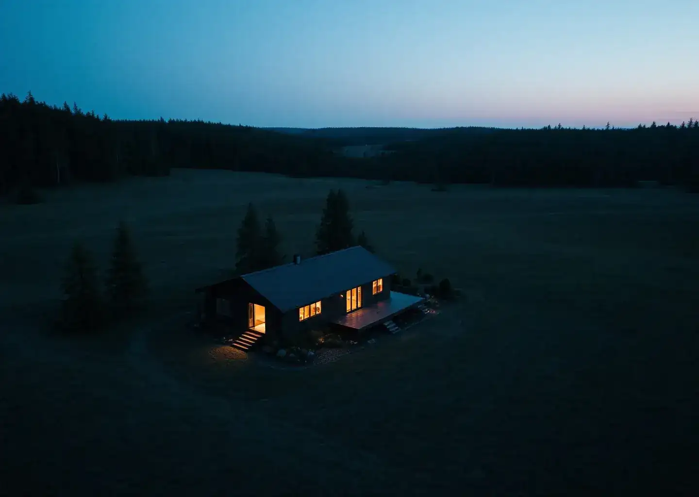 Dusk aerial of a solitary low house with warm lit windows in a dark meadow